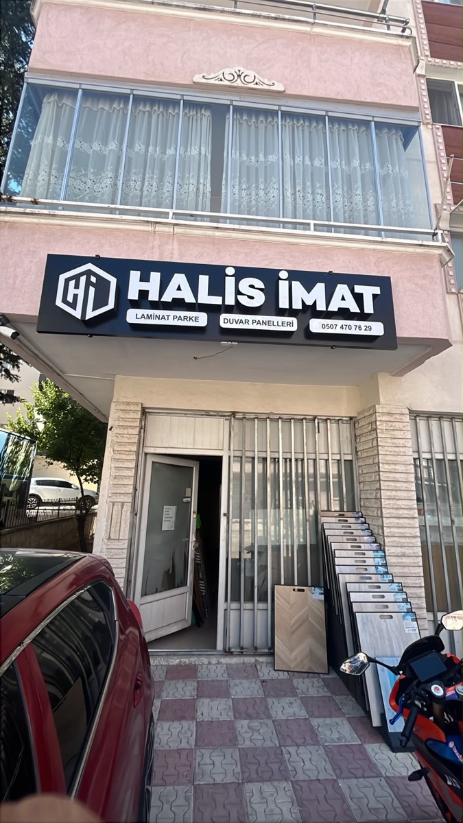 Halis İmat dükkânının sokaktan görünüşü: lacivert tabela ve kapı önünde dizili parke, duvar paneli numuneleri